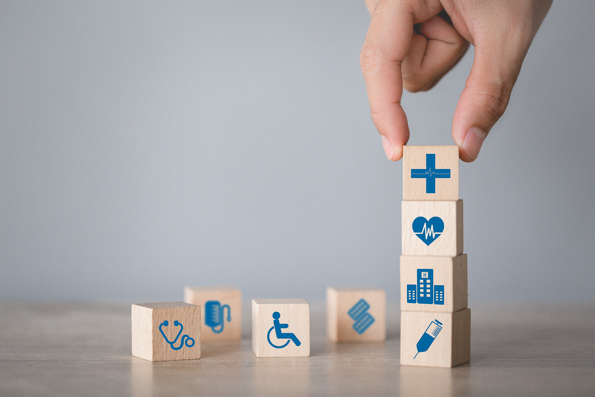 Blocks with health insurance symbols on them. A hand comes from the top to place block on top of stack of blocks.